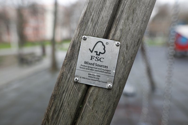 A close-up of a wooden structure with a metal FSC (Forest Stewardship Council) plaque attached, indicating the use of responsibly sourced materials, with a blurred park in the background.